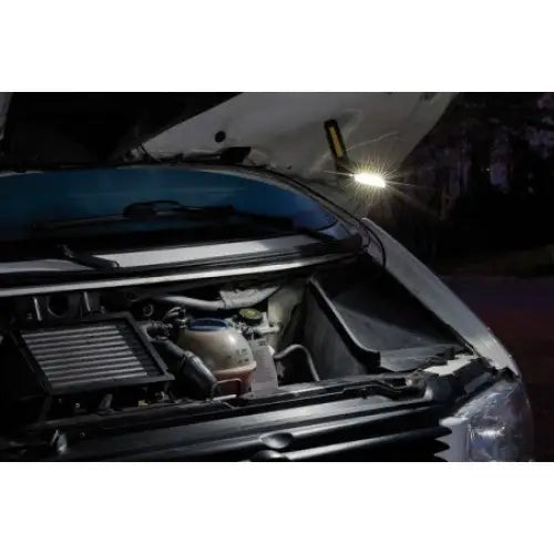 Verkstedslampe LED, montert under panser og lyser motorrommet på bil i mørkt verkstedmiljø.