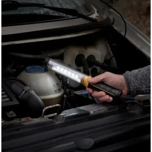 Verkstedslampe med sort og gult håndtak, lyser opp motorrommet mens den holdes i hånden under arbeid.