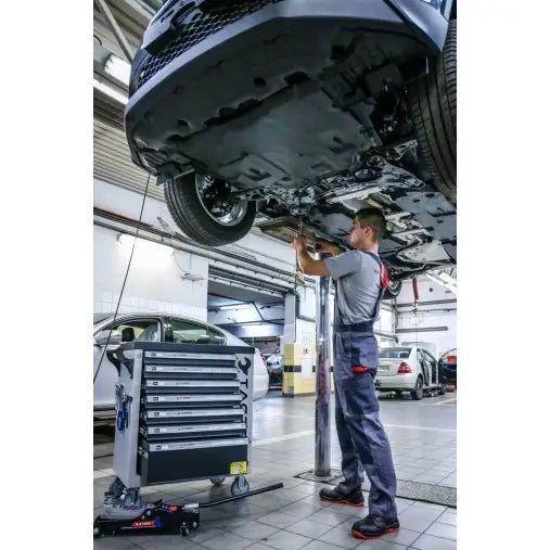 Verkstedvogn Tagred med verktøy, flere skuffer synlige ved bil, mekaniker arbeider under hevet kjøretøy.