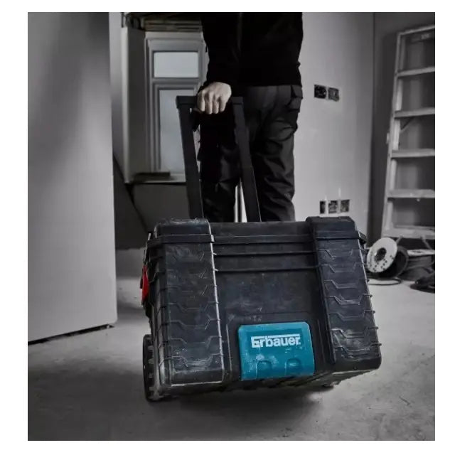 Verktøykasse på hjul, sort og blå modell trilles bortover byggeplass av person i arbeidsklær, sett bakfra.