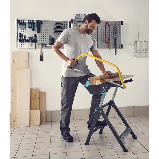Wolfcraft arbeidsbord Master 750 i bruk, sett fra siden mens en treplanke sages med gul håndsag i verksted.