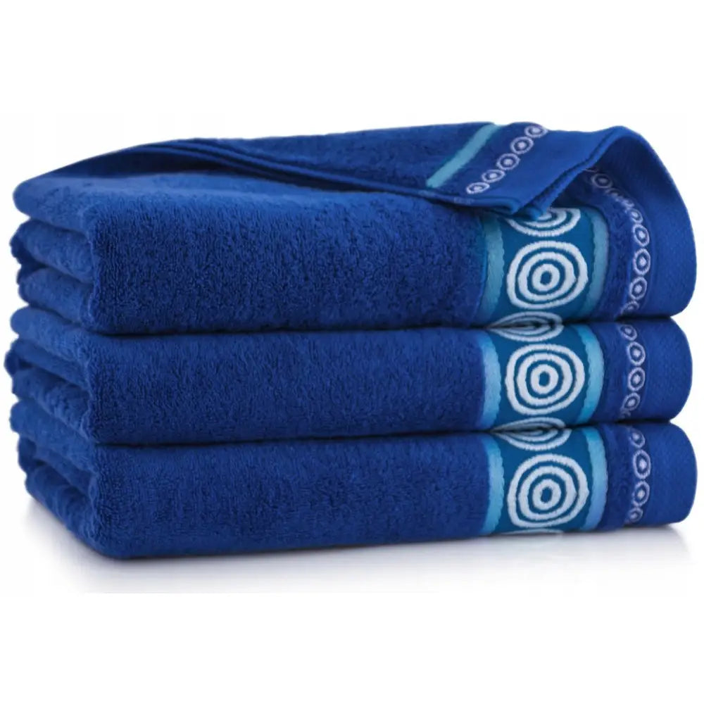 Tre mørkeblå Zwoltex bomullshåndklær, sammenbrettet med dekorative blå og hvite kantbånd, sett fra siden.