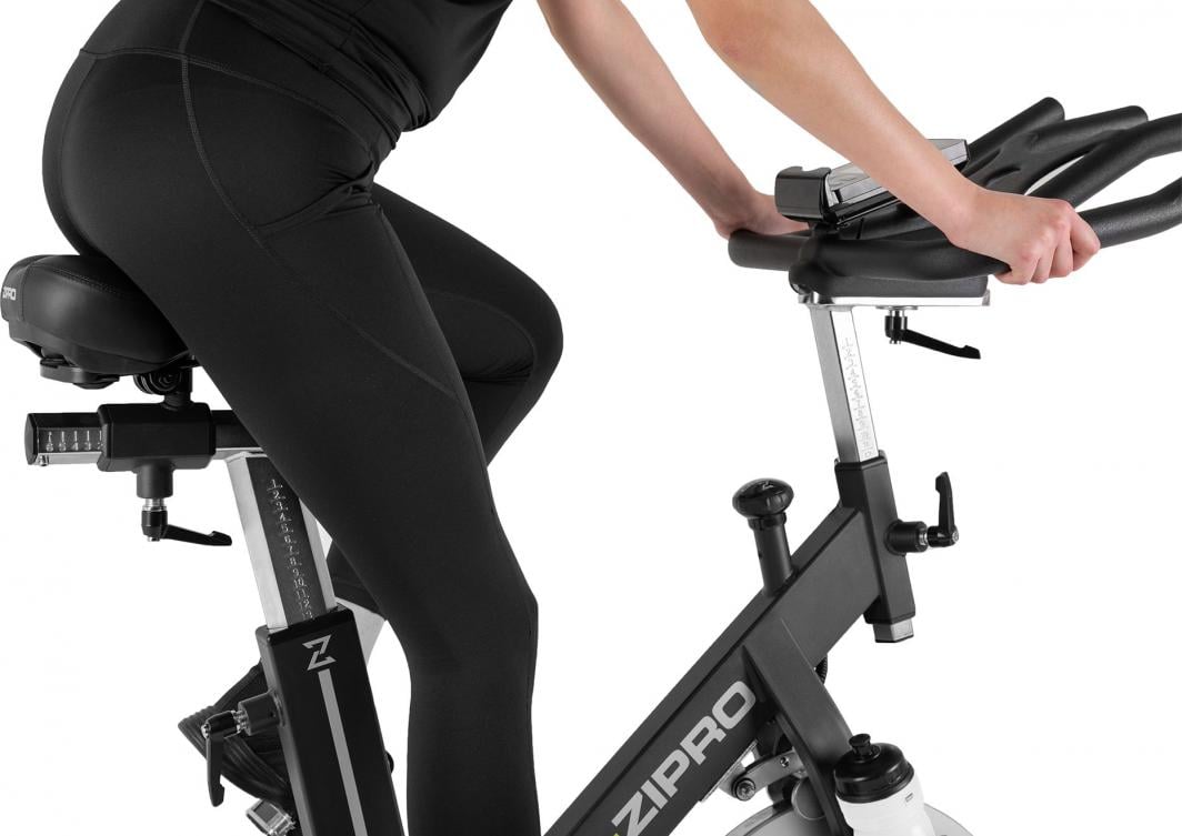 Spinnsykkel i sort og sølv med justerbart sete, en person i treningstøy sykler, sett fra siden.