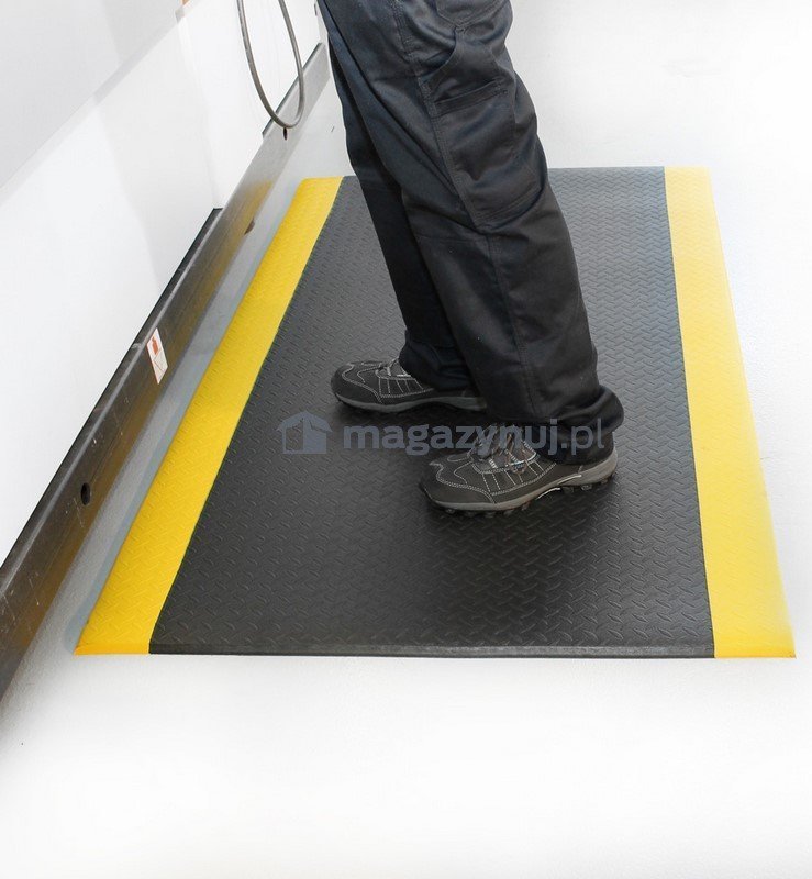 Avlastningsmatte i sort med gul kant på lyst gulv, strukturert overflate, person står på matten med arbeidsbukse og sko.