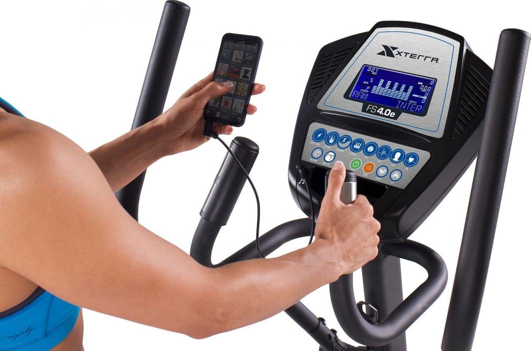 Crosstrainer med digitalt display, flere knapper og mobiltelefon tilkoblet. Person justerer innstillingene.