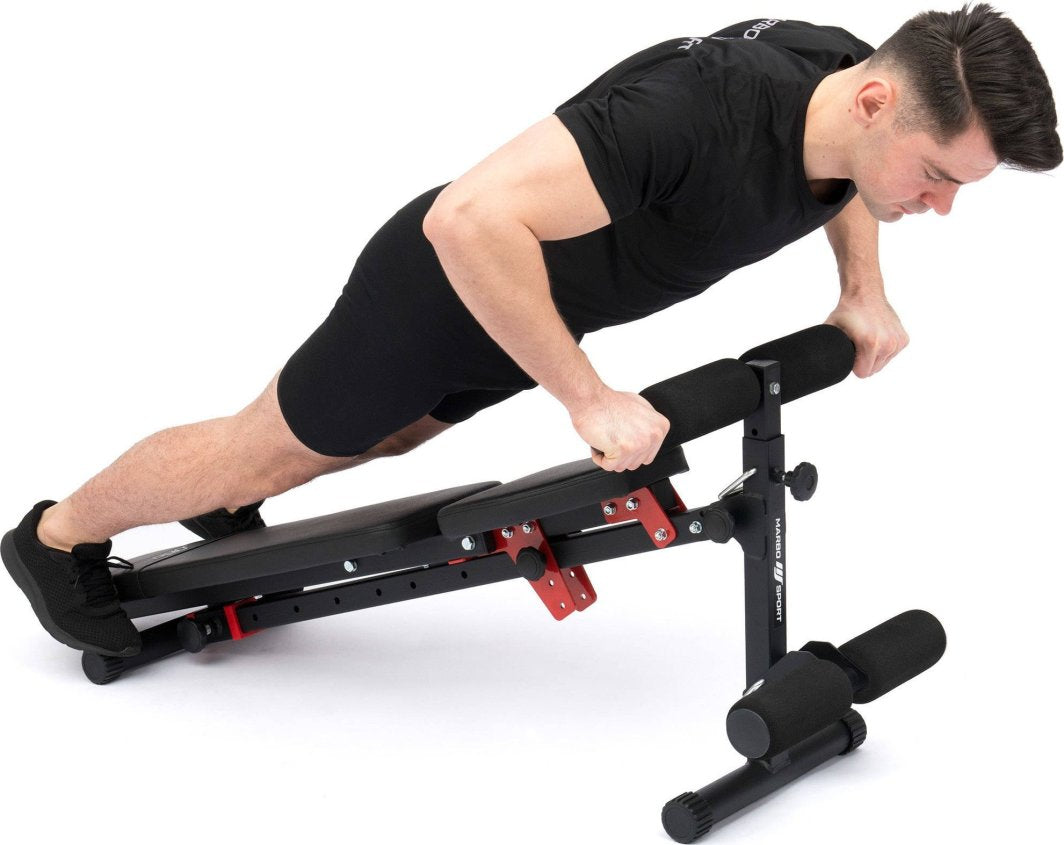 Multifunksjonell Treningsbenk i svart og rødt med polstrede håndtak, brukt av person til styrketrening, sett fra siden.