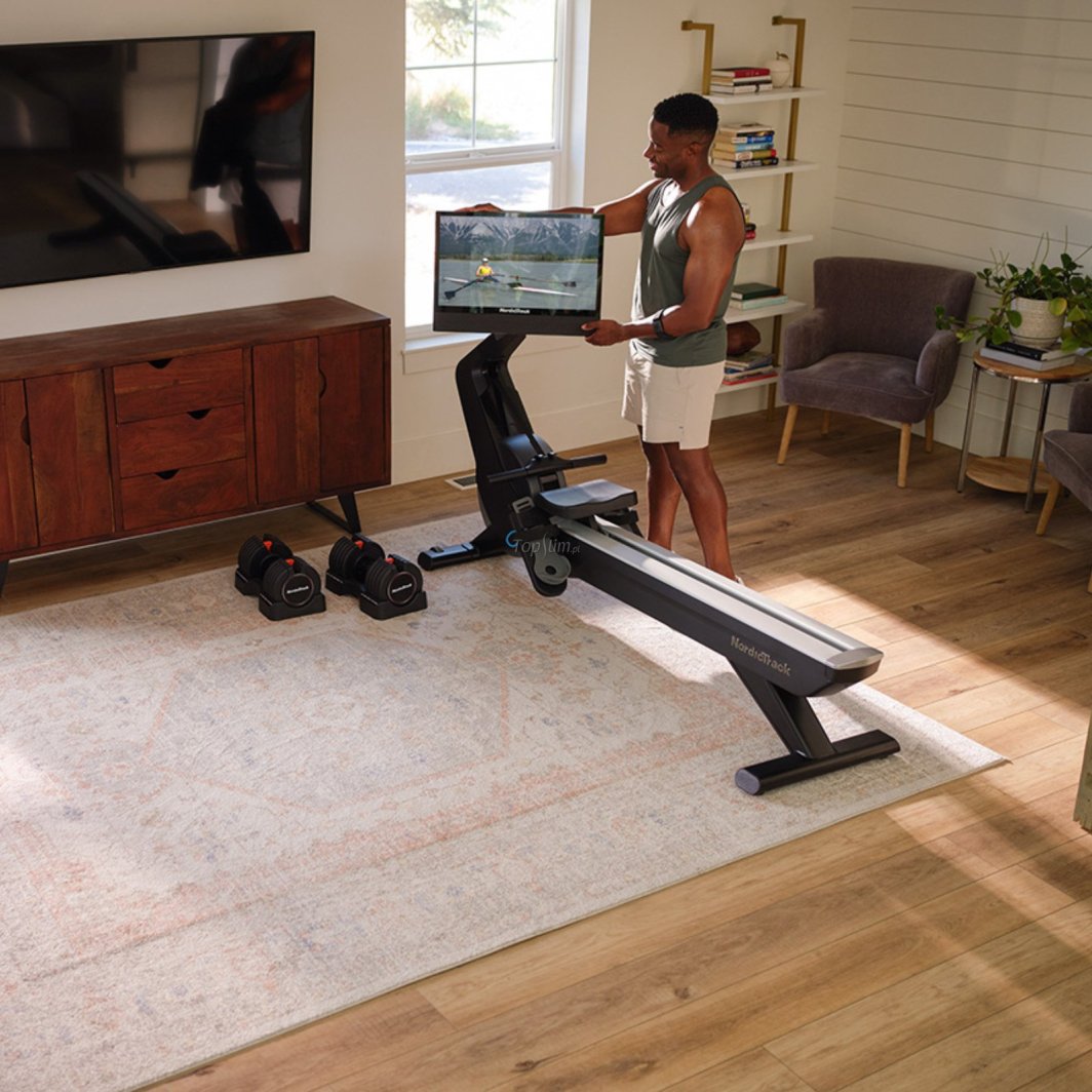 Treningsapparat Hjemme romaskin i sort med stor HD-skjerm, to manualer på gulvet i lyst treningsrom.