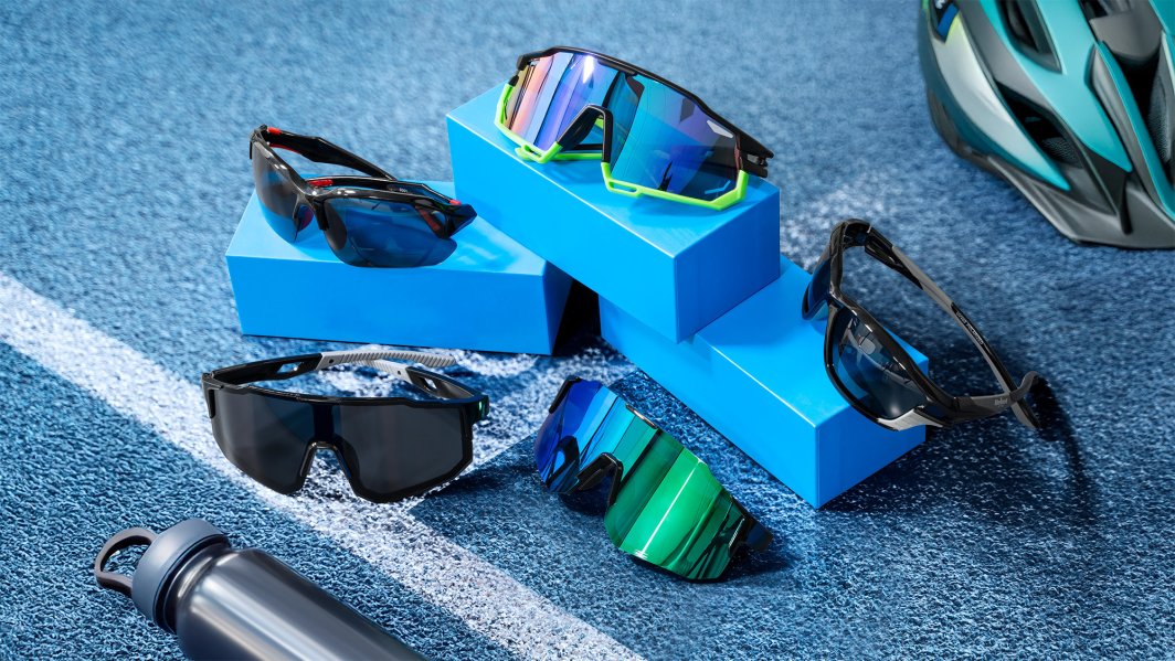 Løpebriller med store glass i fem varianter vises på og rundt blå podiumer, sykkelhjelm og drikkeflaske bak.