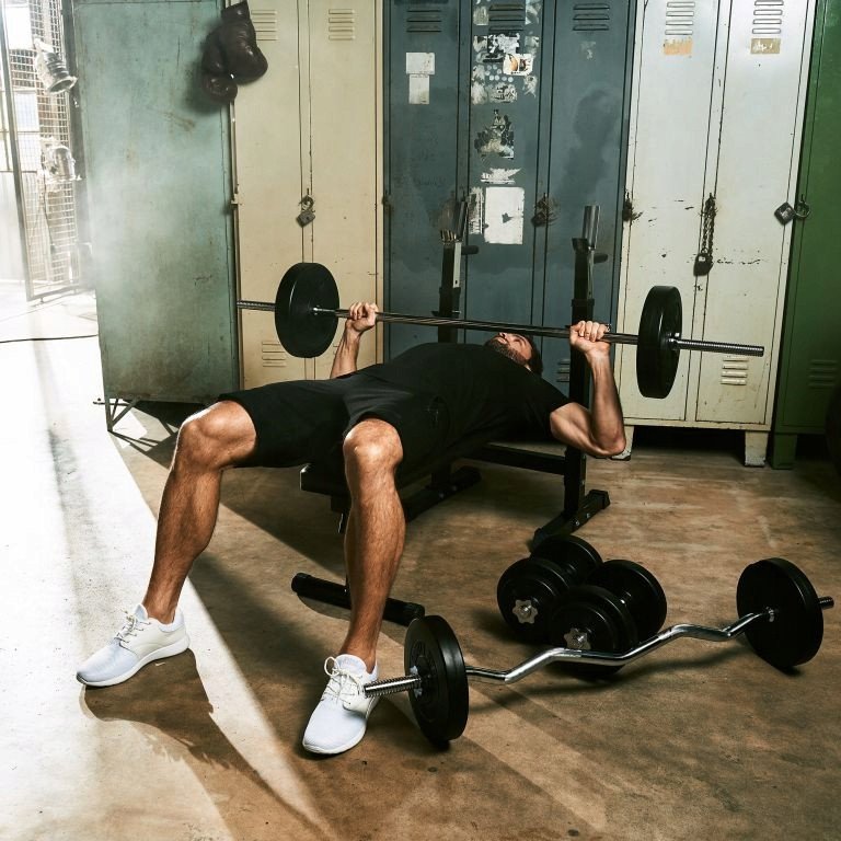 Vektplate på vektstang løftes av person i sort treningstøy på benk, treningsrom med manualer i bakgrunnen.