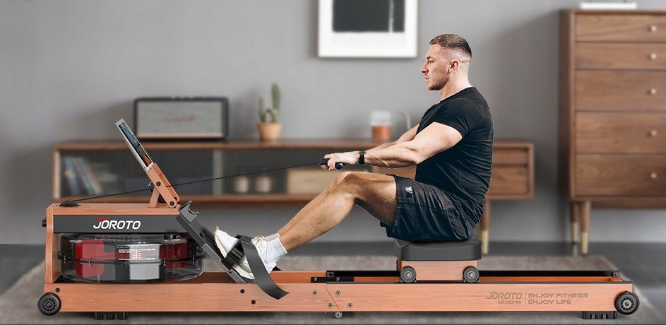 Romaskin i tre med sort sete og fotstøtter, vannbeholder foran, mann trener sittende og drar i håndtaket.