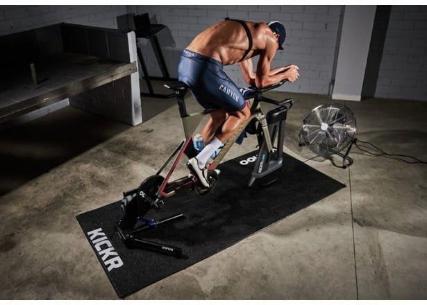 Gymmatte med teksten KICKR under en stasjonær sykkel, mann trener, vifte og utstyr i bakgrunnen.
