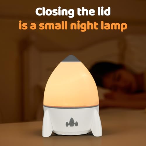 Stjernehimmel Lampe formet som en rakett med lysegul topp, tent på et bord ved siden av en seng.