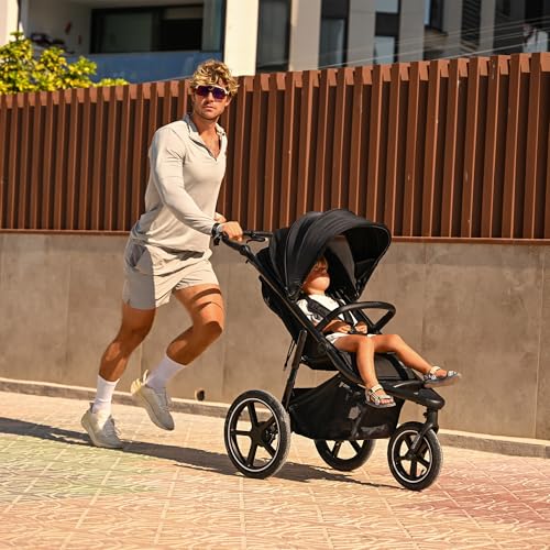 Joggevogn med sort ramme og store hjul, mann jogger på fortau med barn under sort kalesje, sett fra siden.