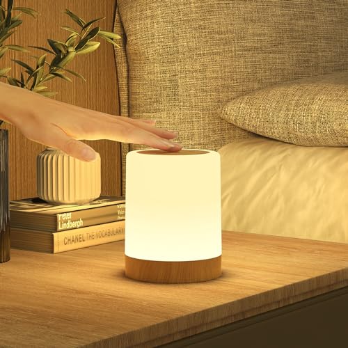 Lampe med hvit sylinderformet skjerm og lys trefarget base, står på nattbord mens en hånd berører toppen.