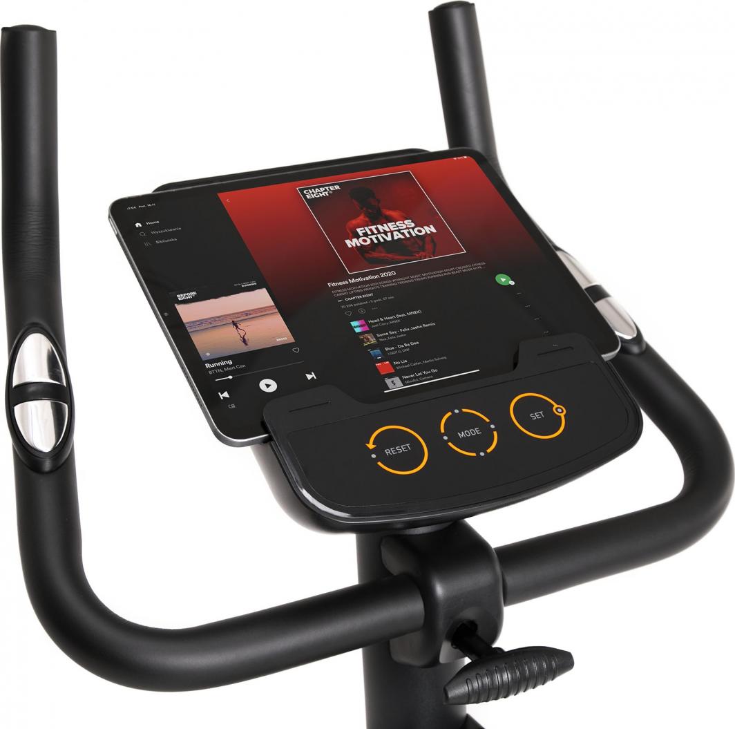 Treningssykkel med sort kontrollpanel, tre oransje knapper og håndtak, tablet med musikk i holderen.