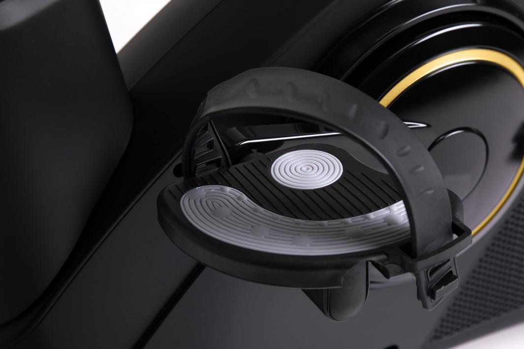 Ergometersykkel med detaljbilde av svart plastpedal, justerbar fotstropp og grått antiskli-felt.