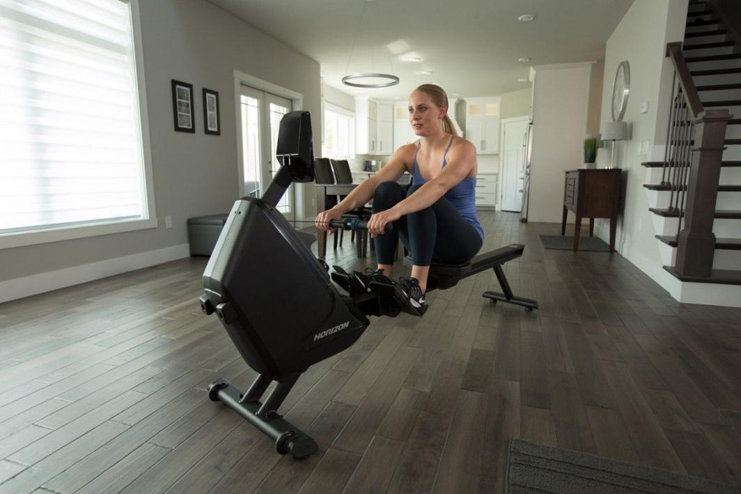 Treningsapparat Hjemme romaskin i sort plast og metall, kvinne trener i lyst, moderne hjemmemiljø.