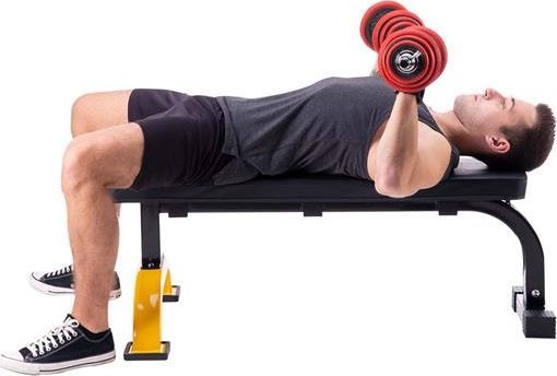 Treningsbenk sett fra siden, med mann i treningsklær som løfter rød manual på sort benk med gule detaljer.