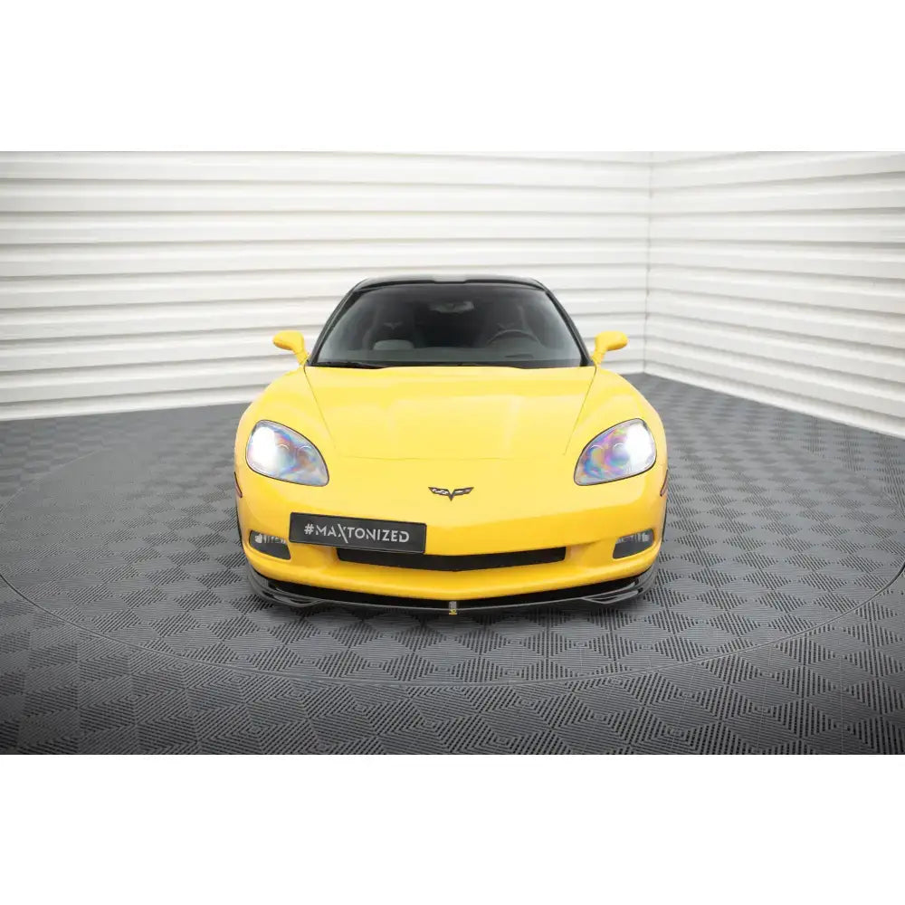 Front lip Chevrolet Corvette C6 i gul, sett rett forfra med svarte detaljer, innendørs på mønstret gulv.