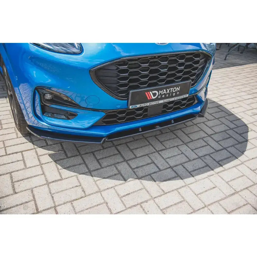 Frontspoiler til Ford Puma St-Line montert på blå bil med sort grill, sett fra lav frontvinkel på brostein.