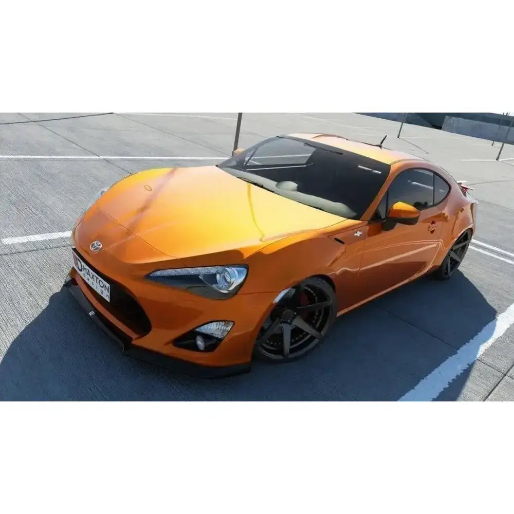 Frontspoiler på oransje Toyota GT86 med sorte felger, sett fra siden og litt forfra på parkeringsplass.