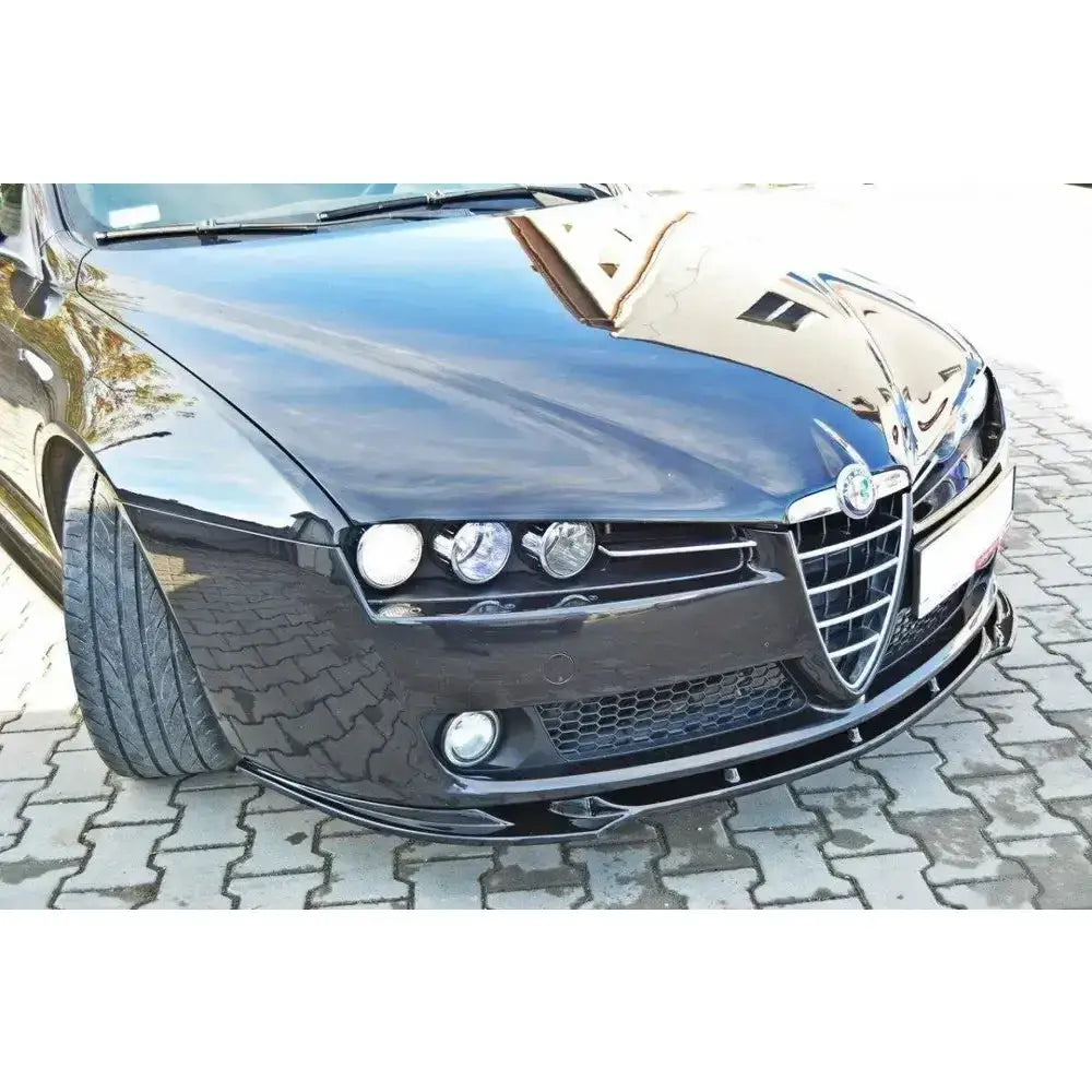 Front lip på Alfa Romeo 159 V.1, sort bil med frontspoiler, grill og lykter på brosteinsbelagt underlag.