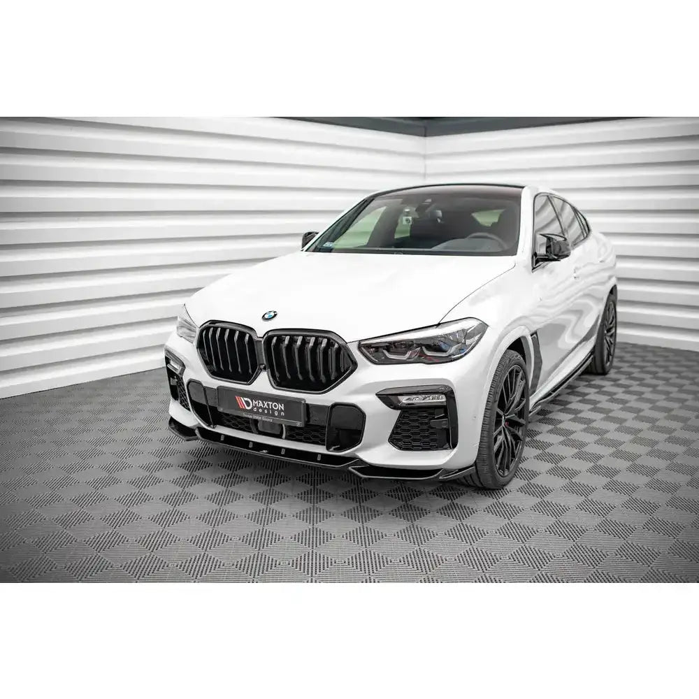 Frontsplitter på hvit BMW X6 M-Pack G06 med sorte detaljer, sett skrått forfra i lyst innendørs lokale.