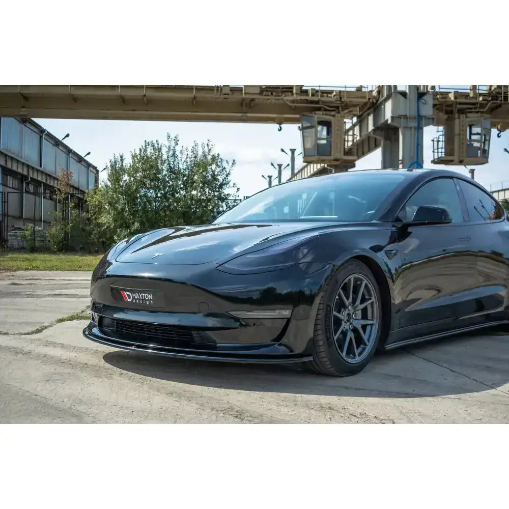 Frontsplitter på Tesla Model 3 V.1 i svart, sett forfra med sportsfelger og industriområde i bakgrunnen.
