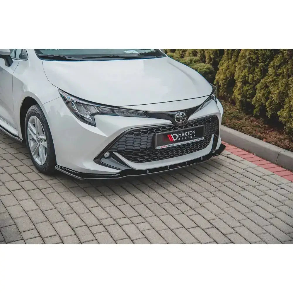 Front lip på hvit Toyota Corolla XII med sort frontspoiler, sett fra skrå vinkel mot steinbelagt bakke.