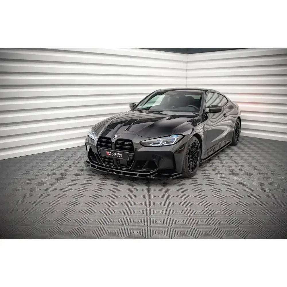Frontspoiler på BMW M4 G82 V.2 i sort høyglans med svarte felger, sett skrått forfra innendørs.