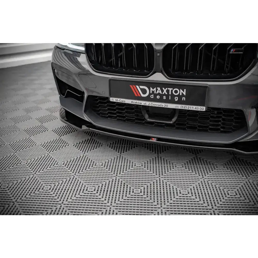 Front lip påmontert BMW M5 F90 Facelift V.2, svart med blank finish, nærbilde forfra mot mønstret gulv.