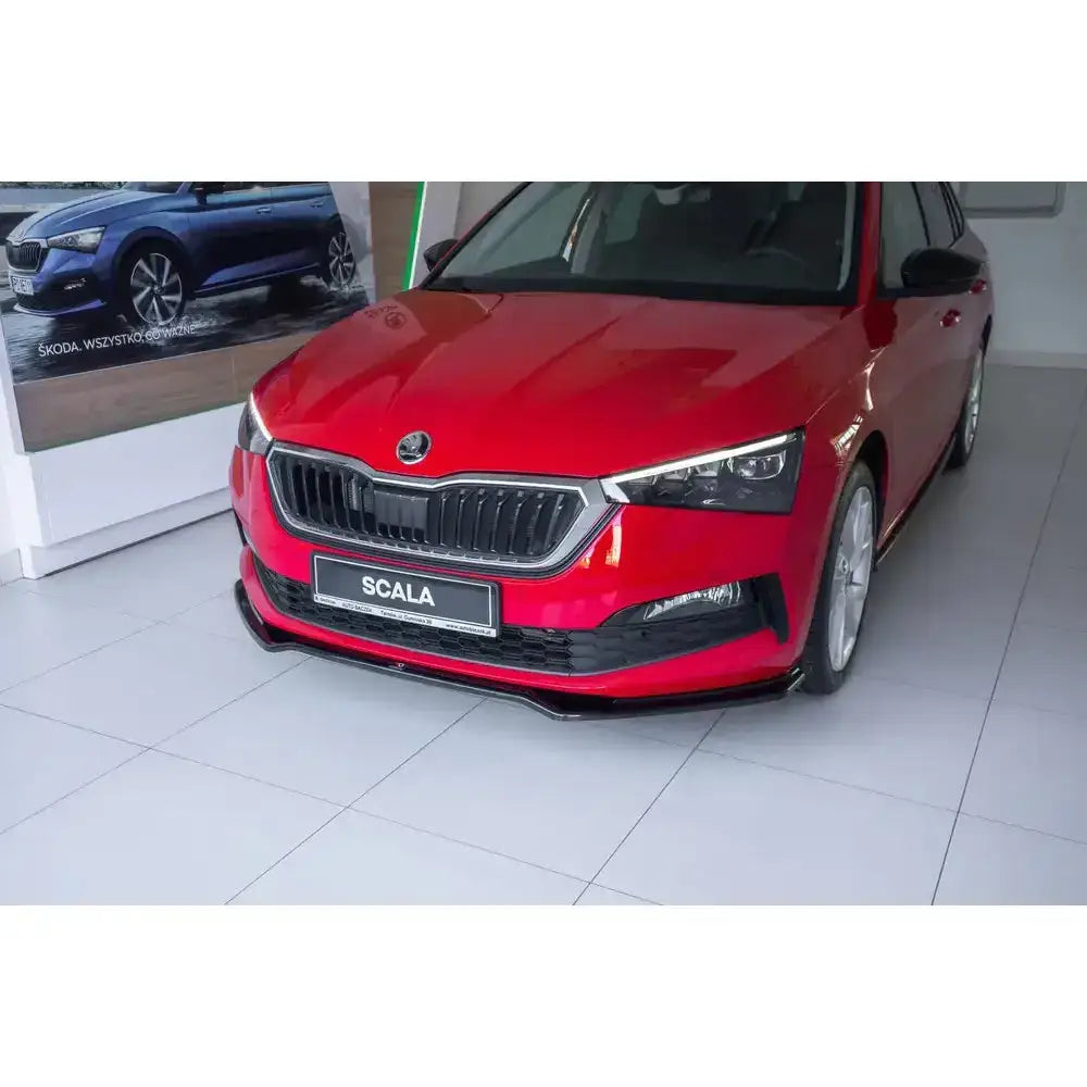 Front lip på rød Skoda Scala V.2 med sorte detaljer, skarpe lykter og blanke felger på flislagt gulv.