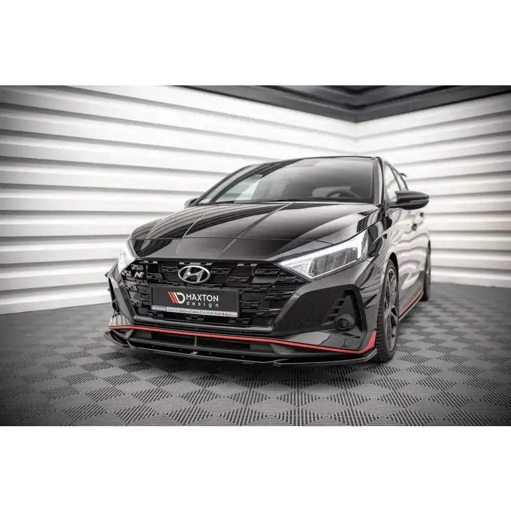 Frontsplitter på sort Hyundai I20 N Mk3 med røde detaljer, sett skrått forfra innendørs.