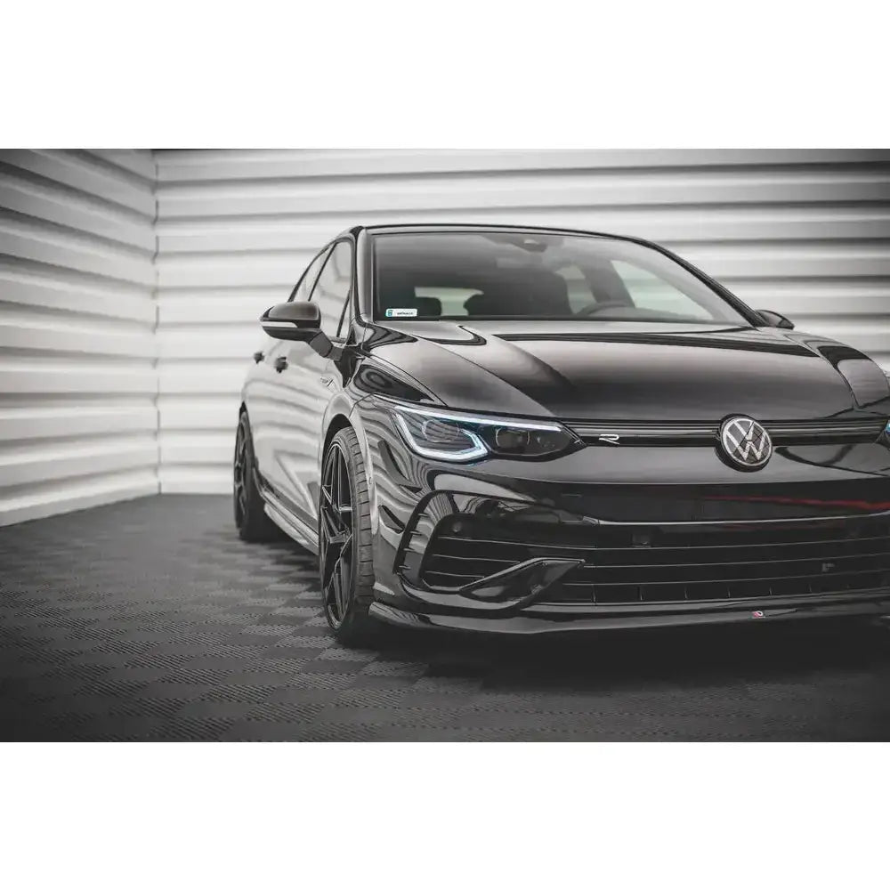 Front lip på Volkswagen Golf R Mk8 V.5 i sort, sett skrått forfra mot lys bakgrunn med metallstruktur.