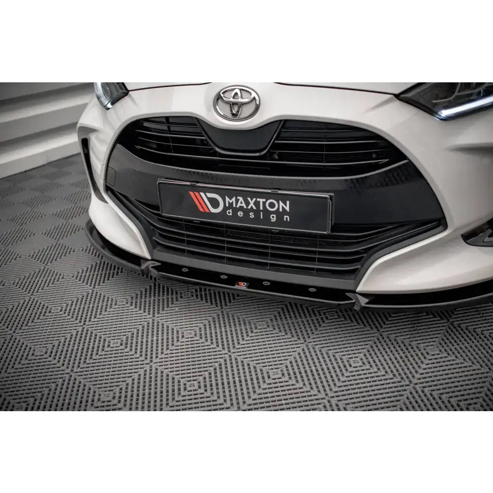 Front lip på Toyota Yaris Mk4 med sort, glanset frontspoiler sett fra skrå vinkel mot geometrisk gulv.