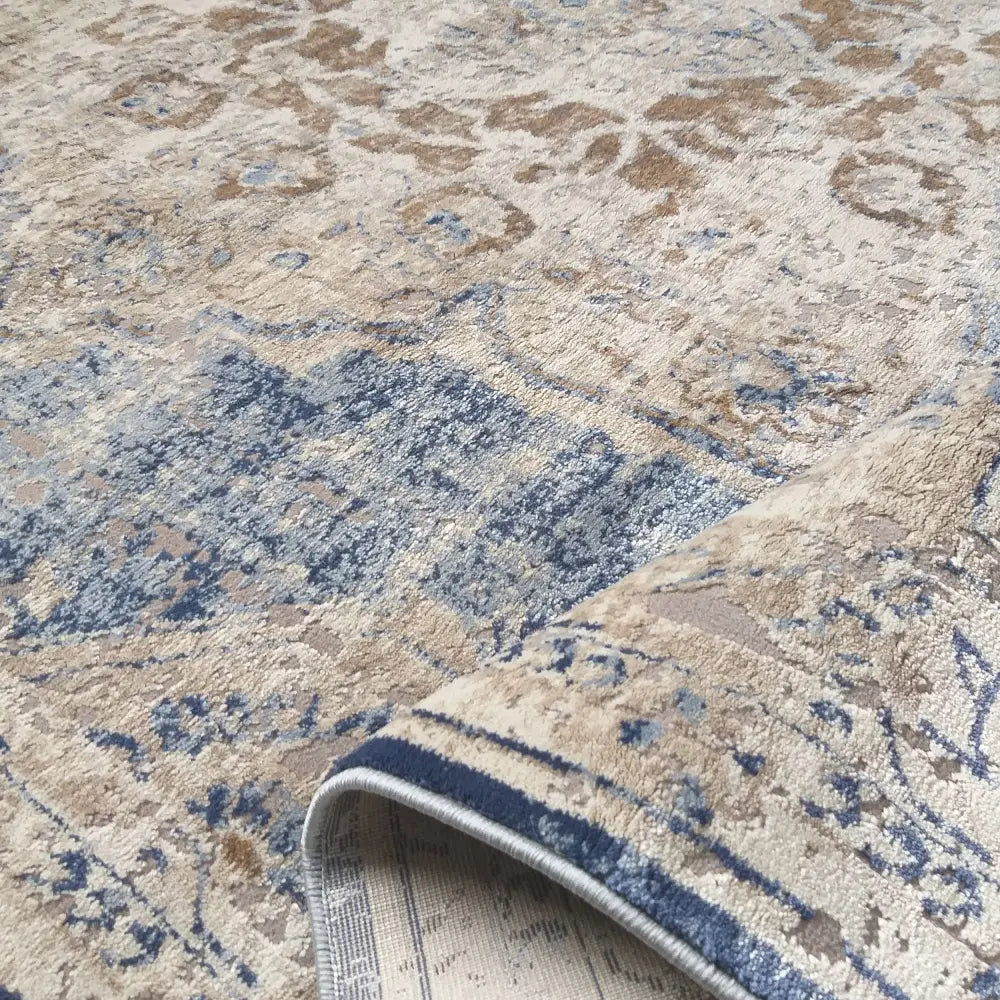 Beige og blått Milas-teppe med orientalsk mønster, skrått vist med oppbrettet hjørne som fremhever teksturen.
