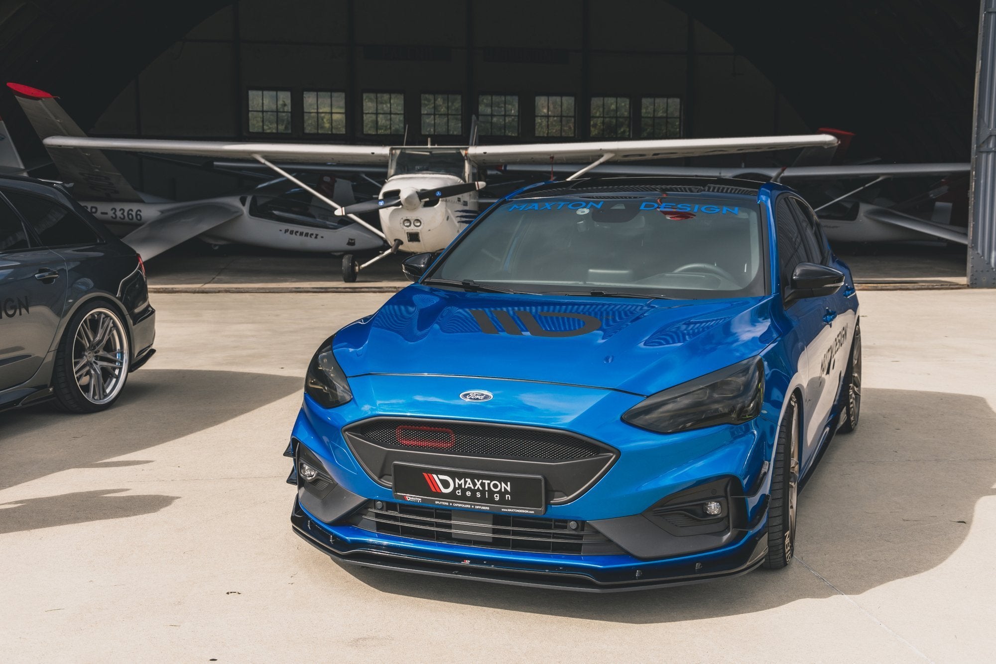 Front lip på Ford Focus MK4 St-Line, blå bil med sorte detaljer foran småfly i hangar.