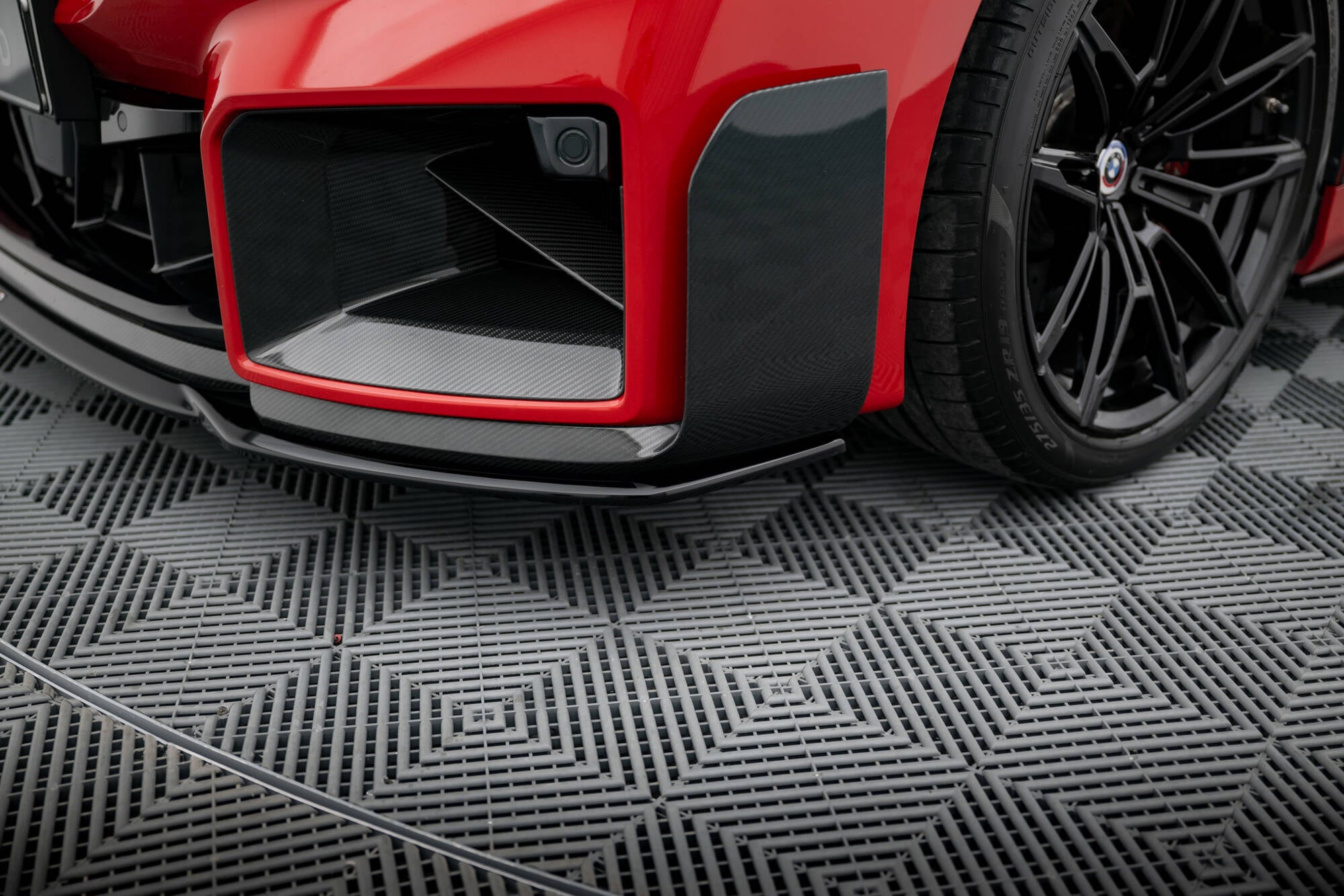 Front lip på BMW M2 M-Performance G87 med karbonfiberdetaljer, rød lakk og synlig felg til høyre.