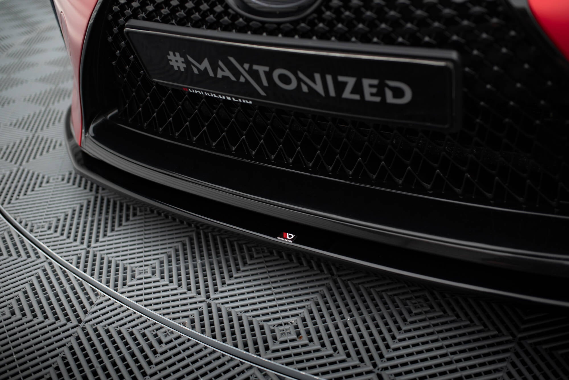 Spoiler Lexus Lc 500 V.2 i sort blank plast montert under bilfanger, nærbilde med logo i midten.