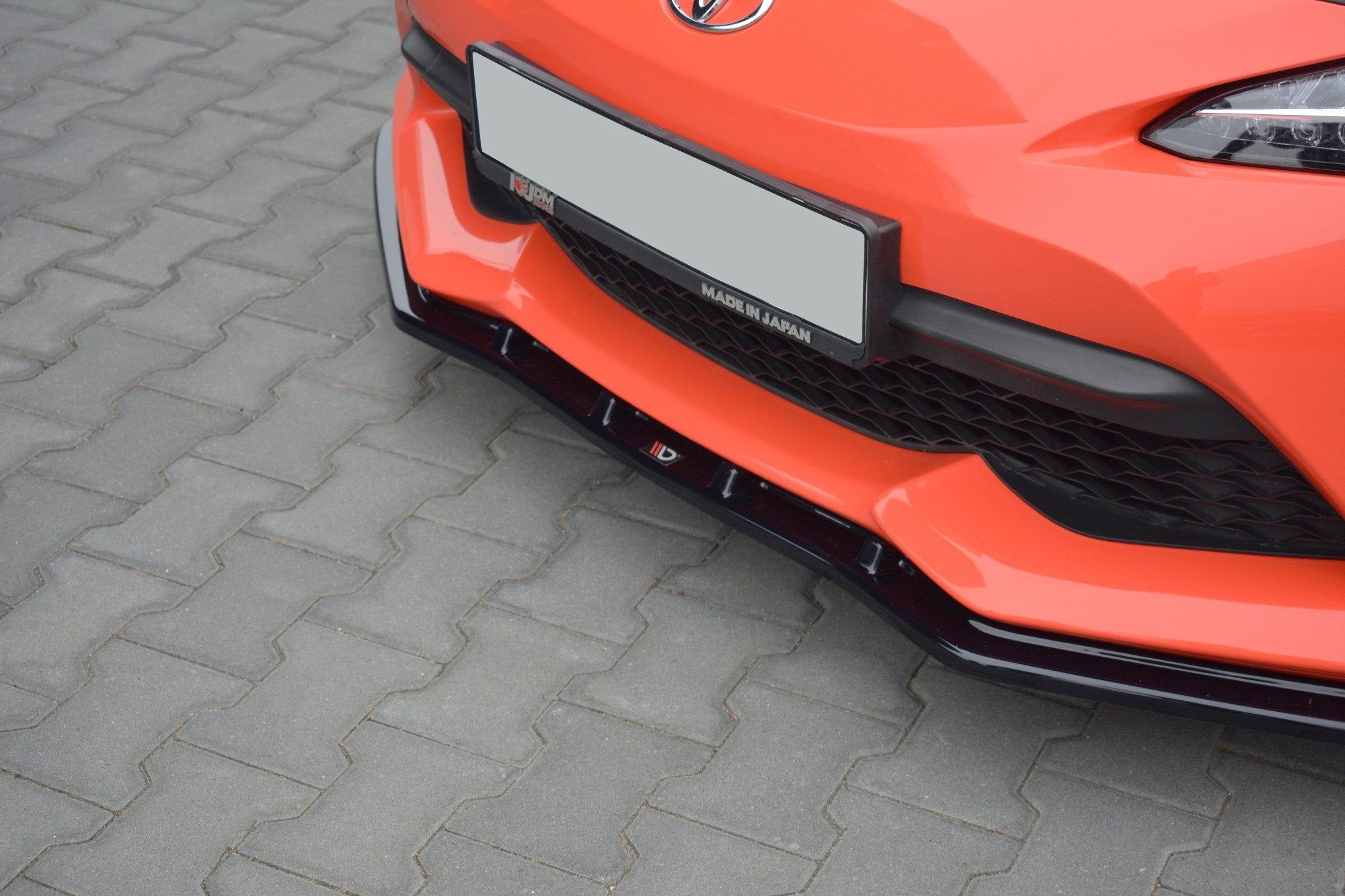 Spoiler Toyota GT86 Facelift V.3 i blank svart på oransje bil, sett skrått forfra med synlig grill.