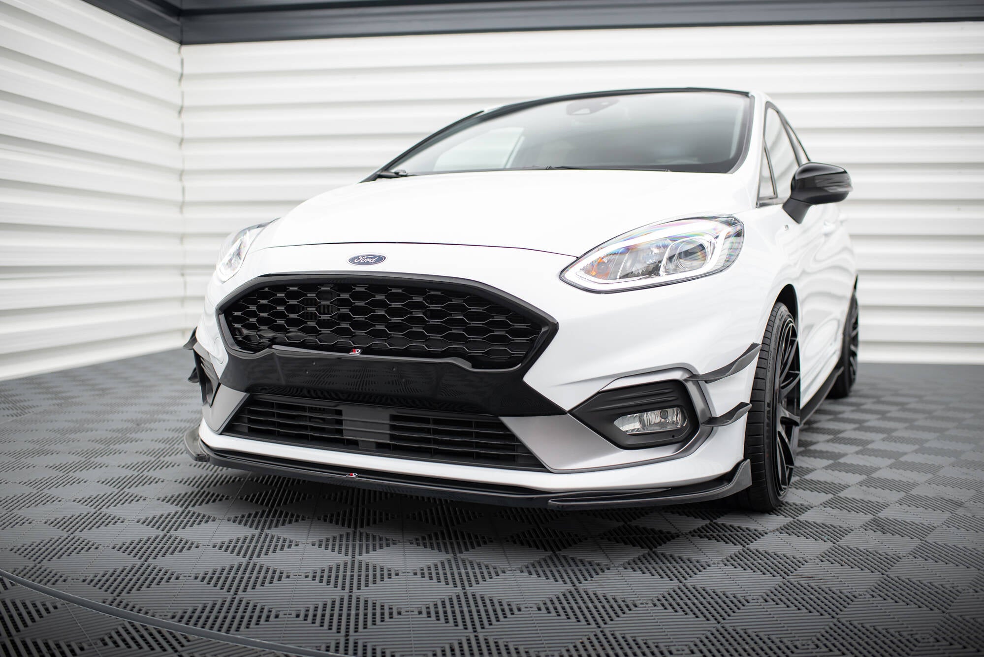 Frontspoiler Ford Fiesta MK8 St i hvitt med sort grill og detaljer, sett skrått forfra i lyst innemiljø.