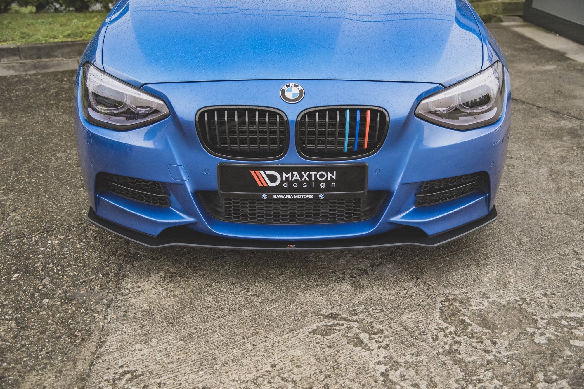 Frontspoiler BMW M135I F20 svart, montert på blå bil med karakteristisk grill og Maxton Design-logo.