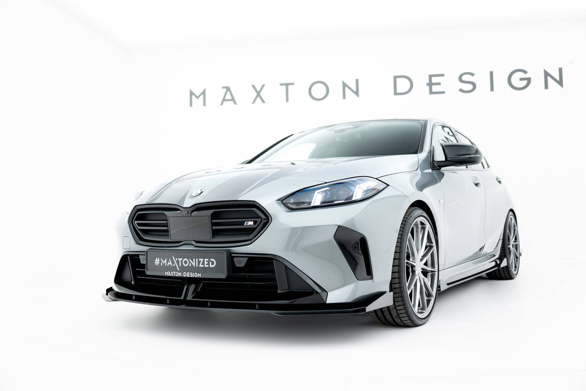 Frontspoiler BMW F70 M135I med flaps V1 montert på lys grå bil med svarte detaljer og Maxton Design-logo.