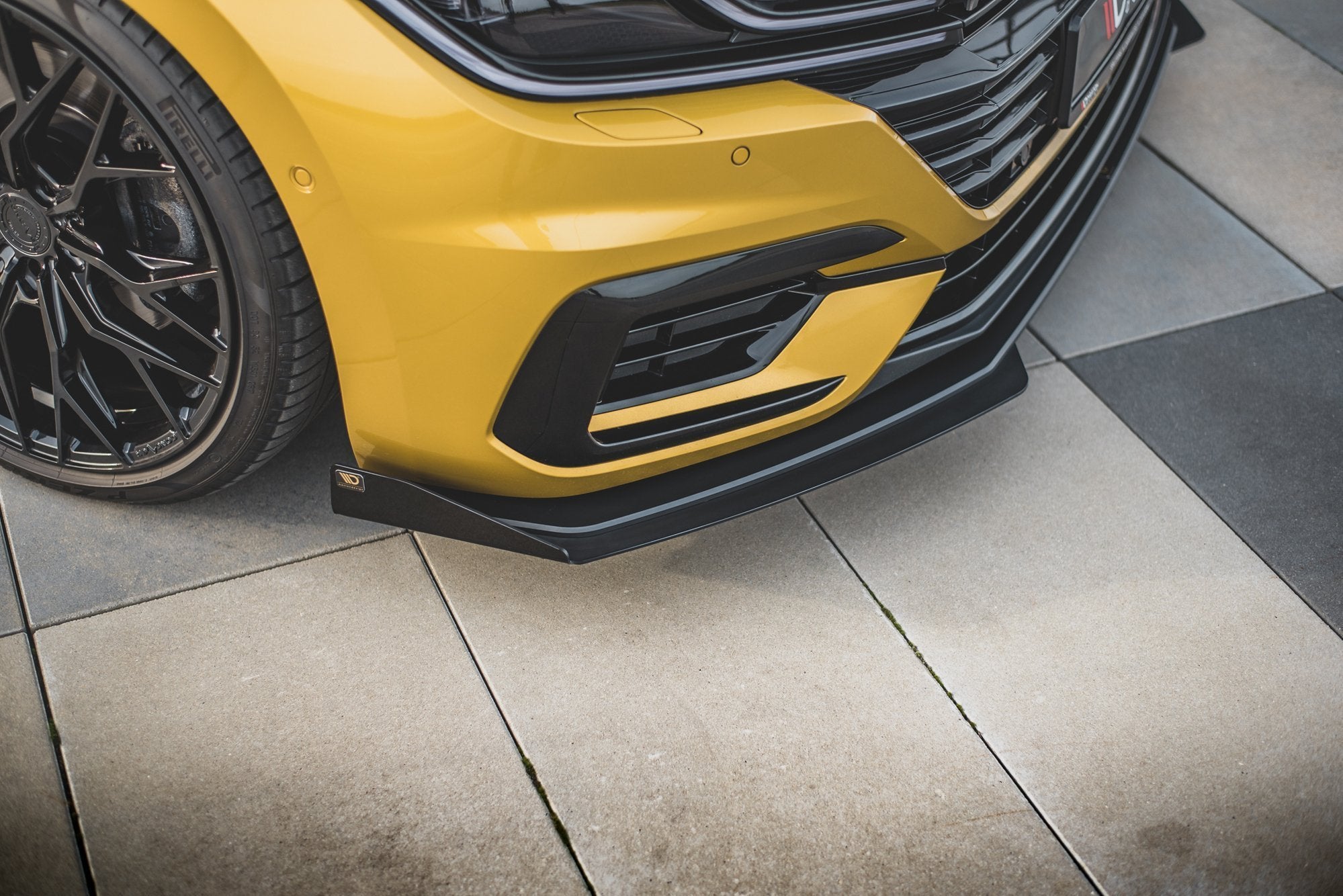 Spoiler til VW Arteon R-Line RS i svart plast under gul støtfanger, med felg og dekk til venstre.