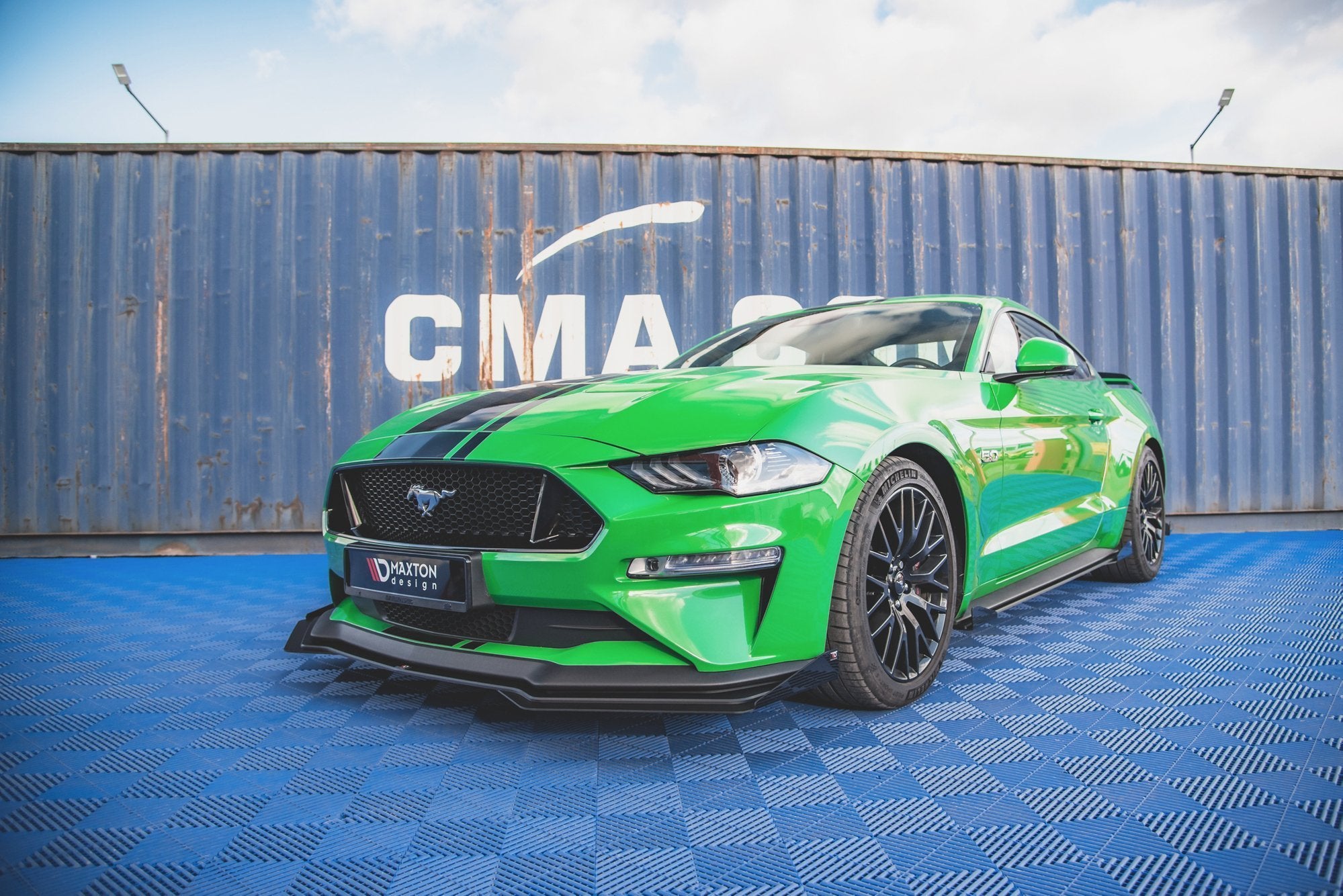 Frontspoiler på grønn Ford Mustang GT MK6 Facelift med svarte felger og racingstriper, blå container bak.