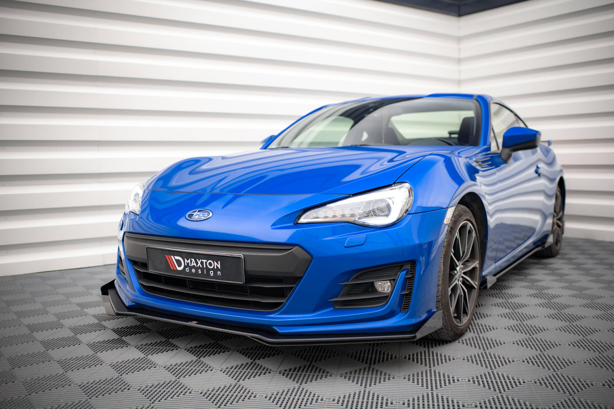Frontspoiler Subaru BRZ MK1 Facelift V.1 montert på blå sportsbil med svarte felger, sett forfra mot hvit vegg.
